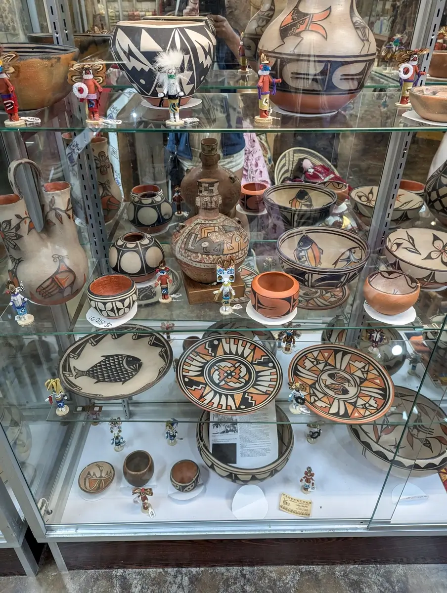 Custer Battlefield Trading Post Cafe Pottery Glass display case filled with handcrafted Native American pottery, bowls, plates, and vases painted with geometric patterns, animals, and earth tone designs. Small figurines are placed among the ceramics, highlighting the variety of traditional styles sold at the trading post.