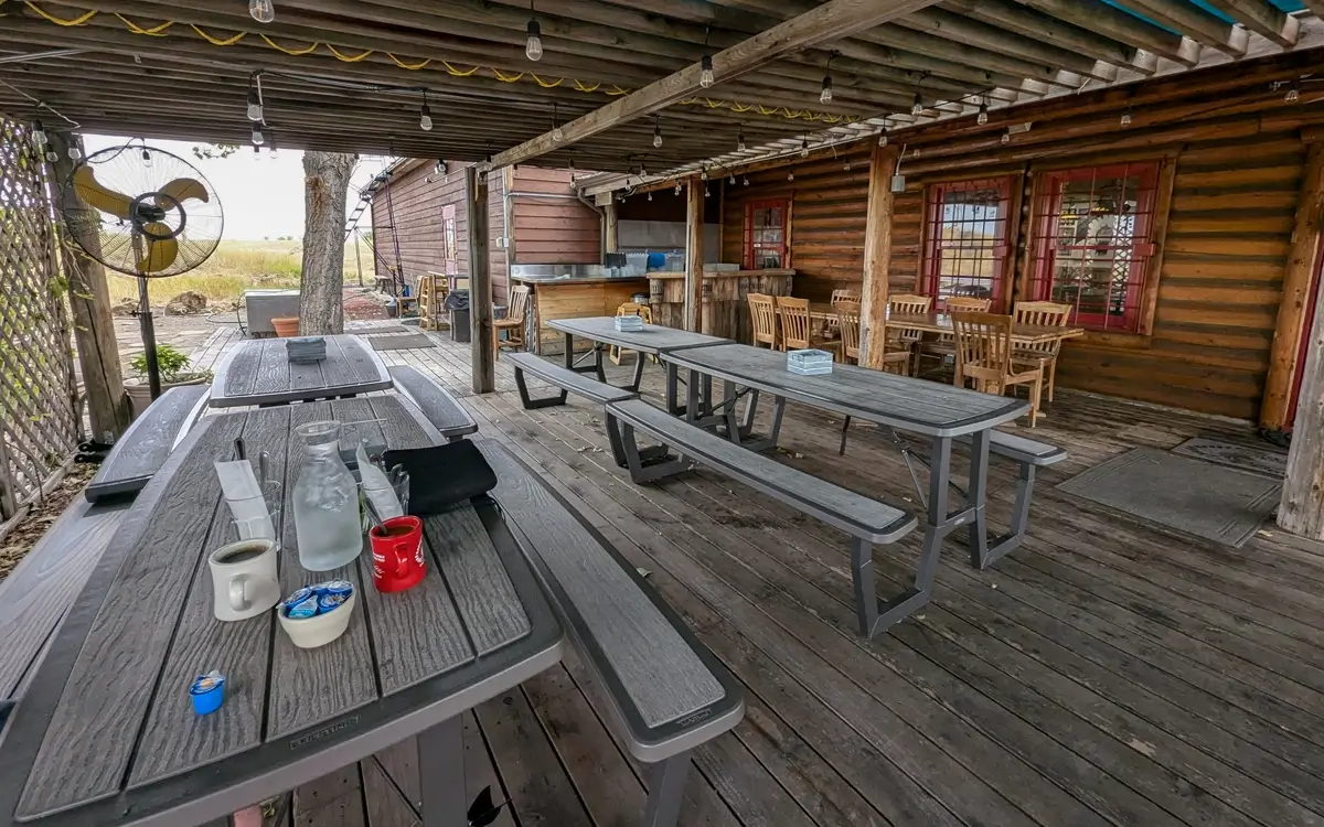 Custer Battlefield Trading Post Cafe Restaurant Covered outdoor patio at Custer Battlefield Trading Post and Cafe with long picnic tables on a wooden deck. Coffee cups, creamers, and a glass bottle sit on one table, while rustic log walls, string lights, and wooden chairs create a relaxed roadside cafe atmosphere.