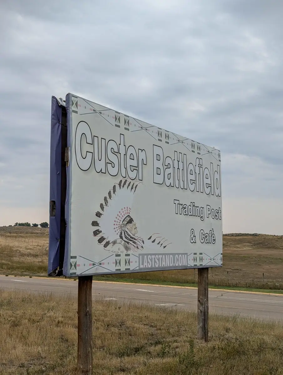 Custer Battlefield Trading Post Cafe Custer Battlefield Trading Post and Cafe roadside sign stands on wooden posts beside a rural highway. The sign shows an illustrated profile of a Native American wearing a feathered headdress and includes the text Custer Battlefield Trading Post and Cafe and laststand.com against an open grassland landscape under a cloudy sky.