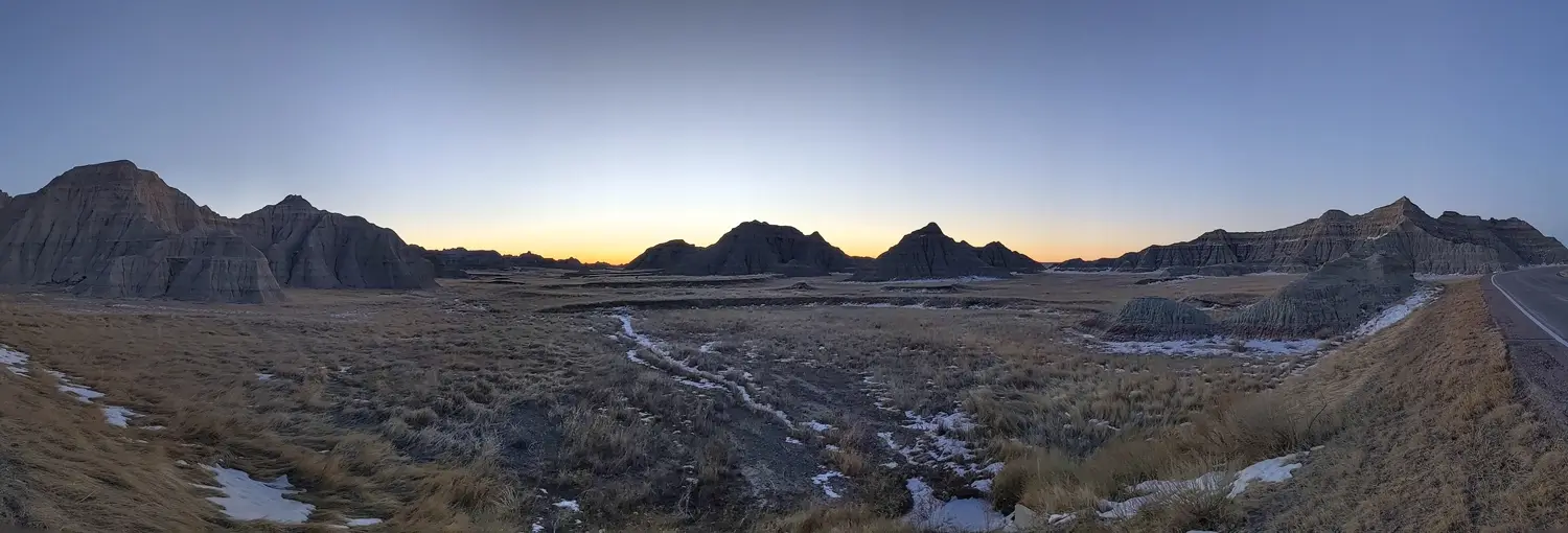 Badlands Sunrise Panorama Wide-angle panorama of a winter sunrise over the jagged peaks and prairie of Badlands National Park, featuring a golden horizon and snow-covered ground.