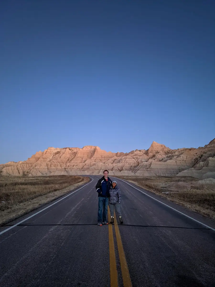 Minerva And Sam In The Badlands A couple standing in the middle of a deserted paved road winding through the towering rock formations of the Badlands at dusk.