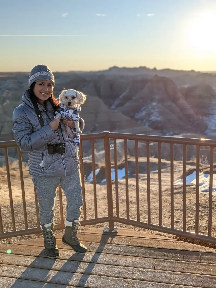 Minerva And Toby Badlands Portait Portrait of a woman holding a small white dog in a sweater at a scenic overlook in the Badlands, with the sun setting behind them.