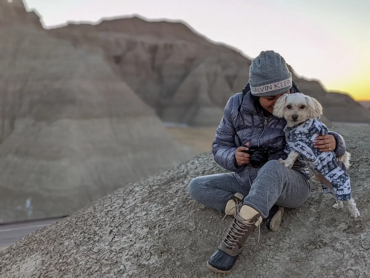 Minerva And Toby In Badlands A woman sitting on a grey clay mound hugging her small dog while visiting the rugged terrain of Badlands National Park in winter.