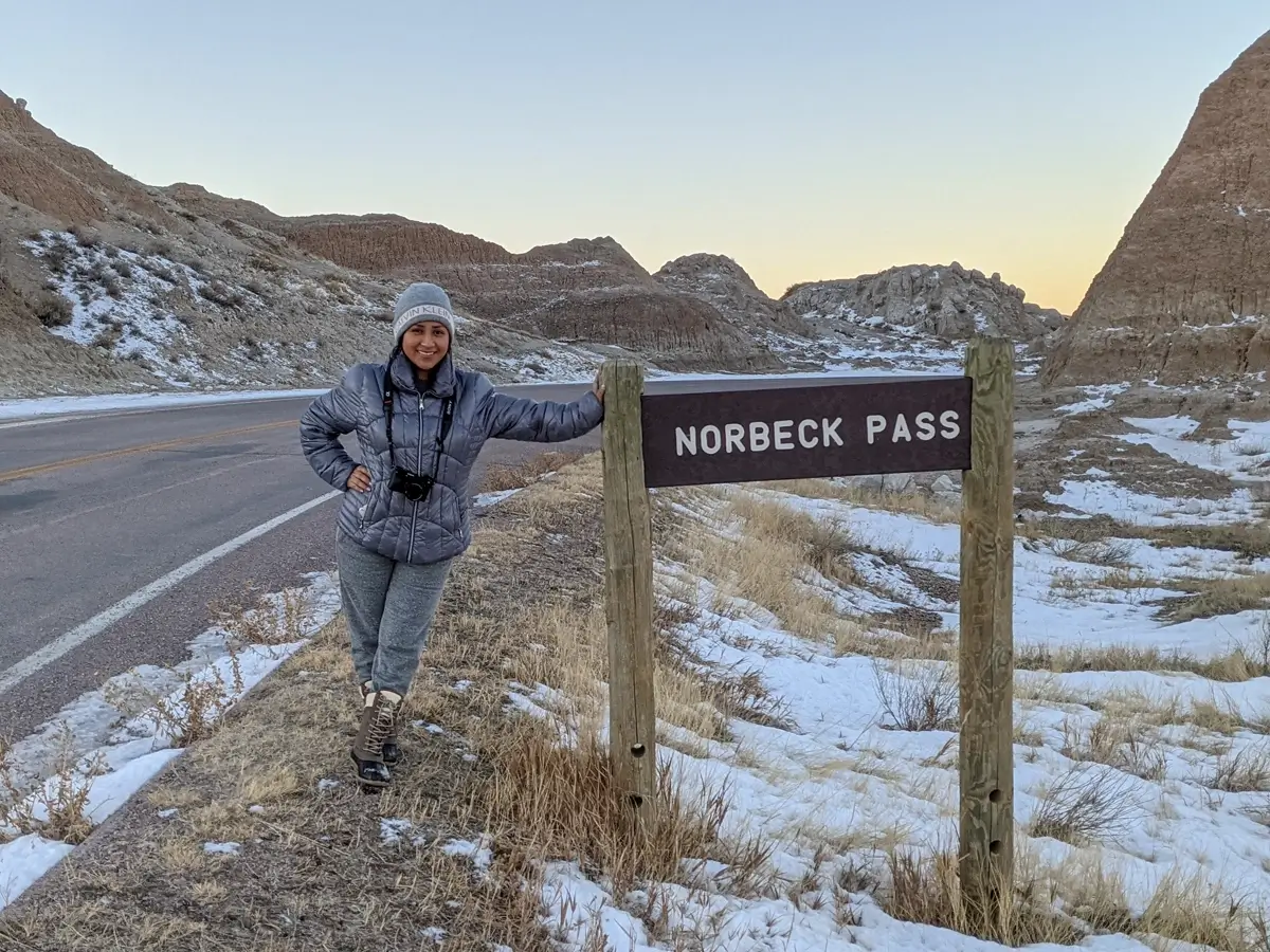 Minerva At Norbeck Pass A woman in a puffer jacket and winter gear standing next to the Norbeck Pass elevation sign on a snowy road in Badlands National Park.