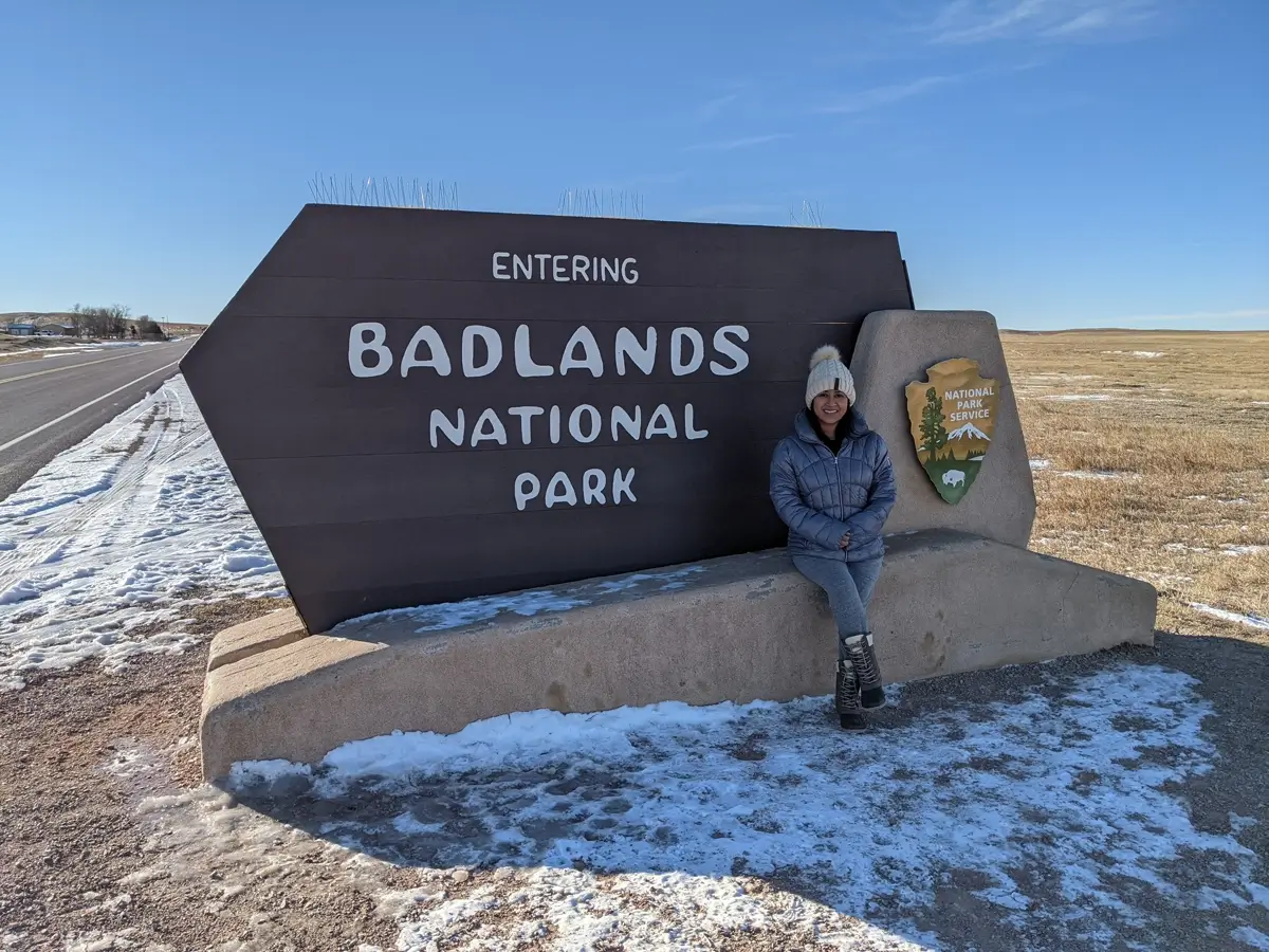 Minerva At The Badlands NP Sign A traveler sitting on the iconic wood and stone entrance sign for Badlands National Park, South Dakota, surrounded by winter prairie.