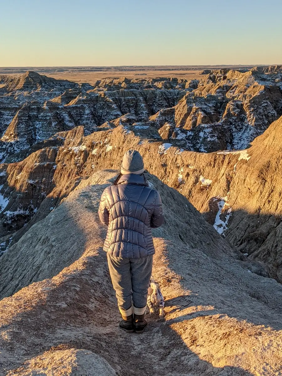 Minerva With Snowy Badlands A woman looking out over a vast valley of snow-dusted ridges and spires from a high viewpoint in Badlands National Park at sunset.