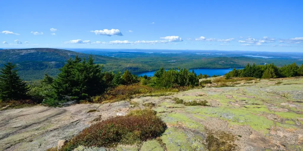 A panoramic view from a rocky overlook showing forests, rolling hills, and a blue lake below. The scene conveys elevation, natural wilderness, and expansive outdoor scenery on a clear day. Credit to LMMiller9 on Pixabay.