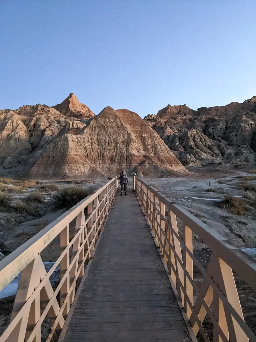 Saddle Pass Trail Wooden boardwalk leading toward layered rock formations in Badlands National Park during winter, with a person standing at the center and soft golden light illuminating the rugged cliffs. The barren landscape and pale blue sky highlight the dramatic textures of the Badlands in South Dakota.