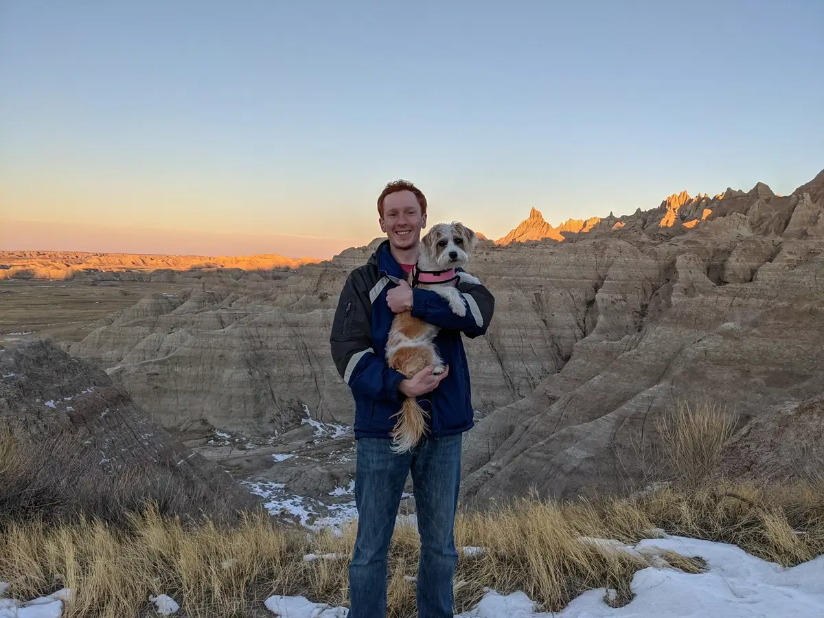 Sam And Lily Badlands A man holding a small scruffy dog in a pink harness standing in front of the golden-hour light illuminating the Badlands peaks in winter.