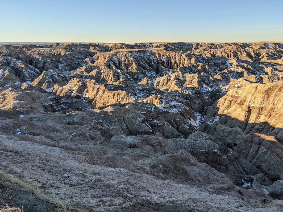 Snowy Badlands National Park High-angle view of the rugged white and tan peaks of Badlands National Park covered in a light blanket of winter snow under a clear sky.