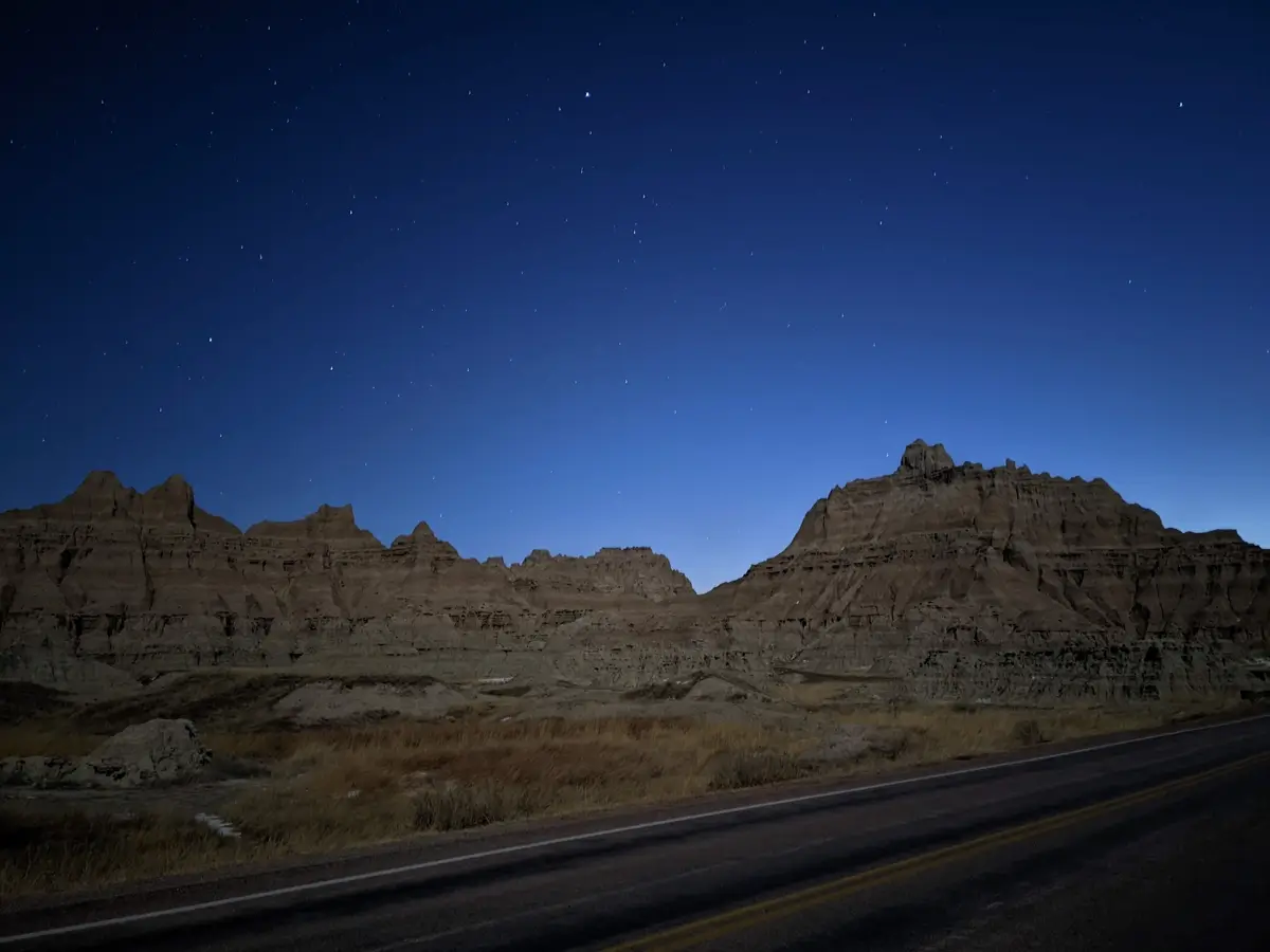 Stars In The Badlands Starry night sky above jagged Badlands rock formations, with a quiet road running along the base of the cliffs in winter. The deep blue twilight and scattered stars create a peaceful nighttime scene in Badlands National Park, South Dakota.