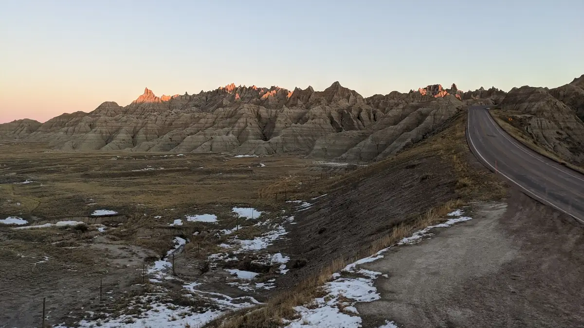 Sunrise In The Badlands Early morning light hitting the jagged spires of the Badlands Loop Road in South Dakota, featuring snowy patches and a pink horizon.
