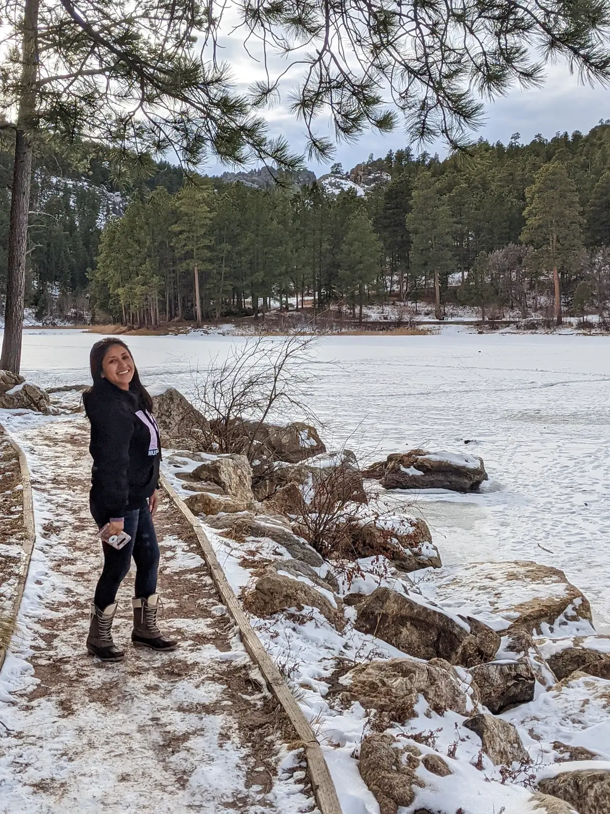 A woman smiling while walking on a snowy path with rock borders alongside frozen Horse Thief Lake in the Black Hills.