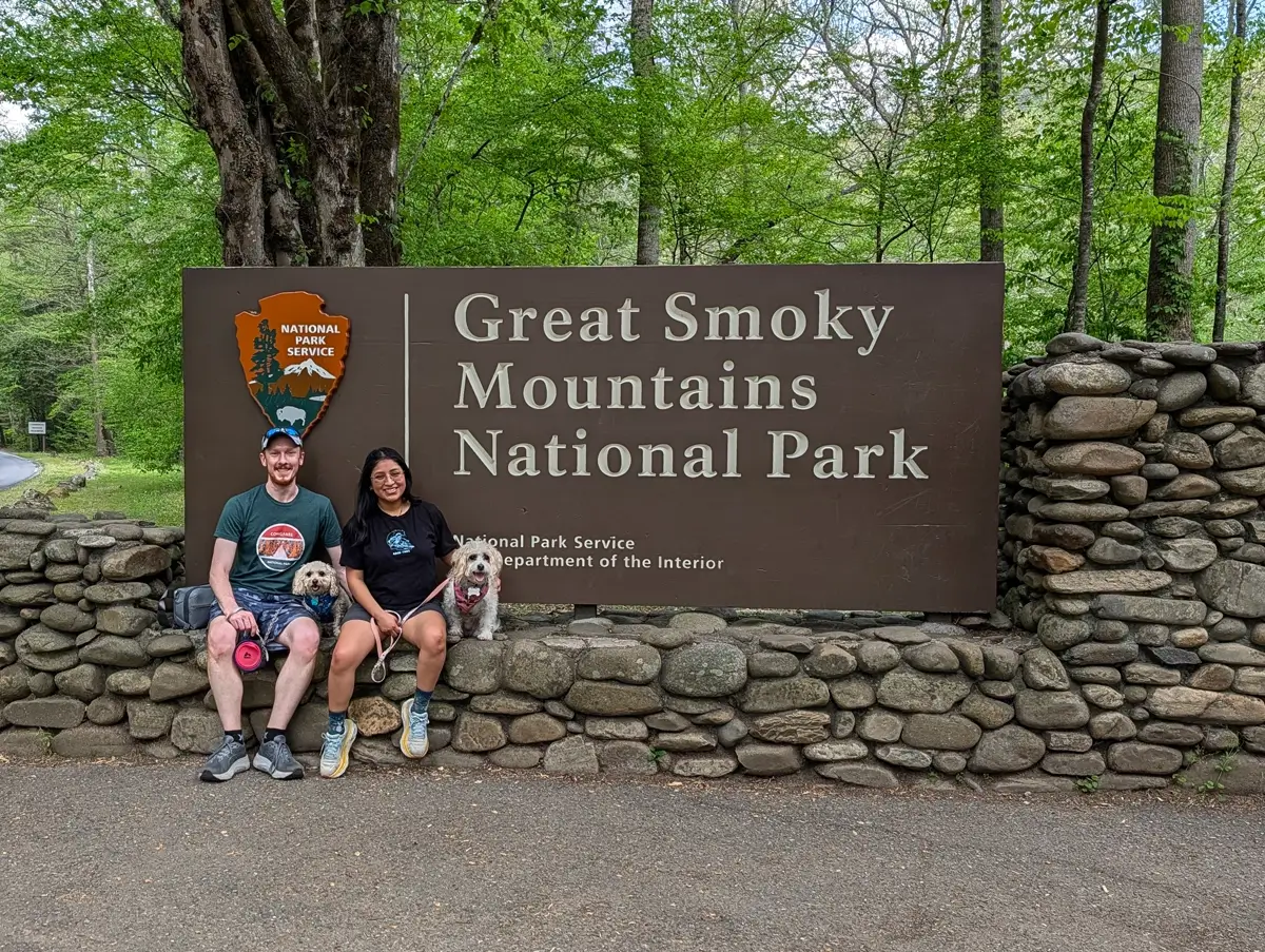 A man and woman sitting on a stone wall in front of the Great Smoky Mountains National Park entrance sign with their two small dogs, Toby and Lily.