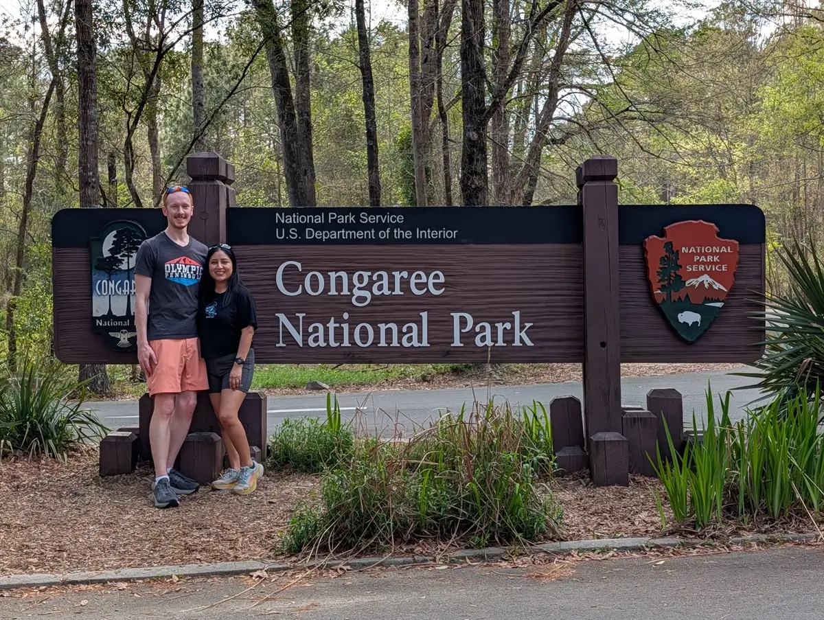 A couple poses proudly in front of the large wooden Congaree National Park sign, a landmark for hikers visiting the area.