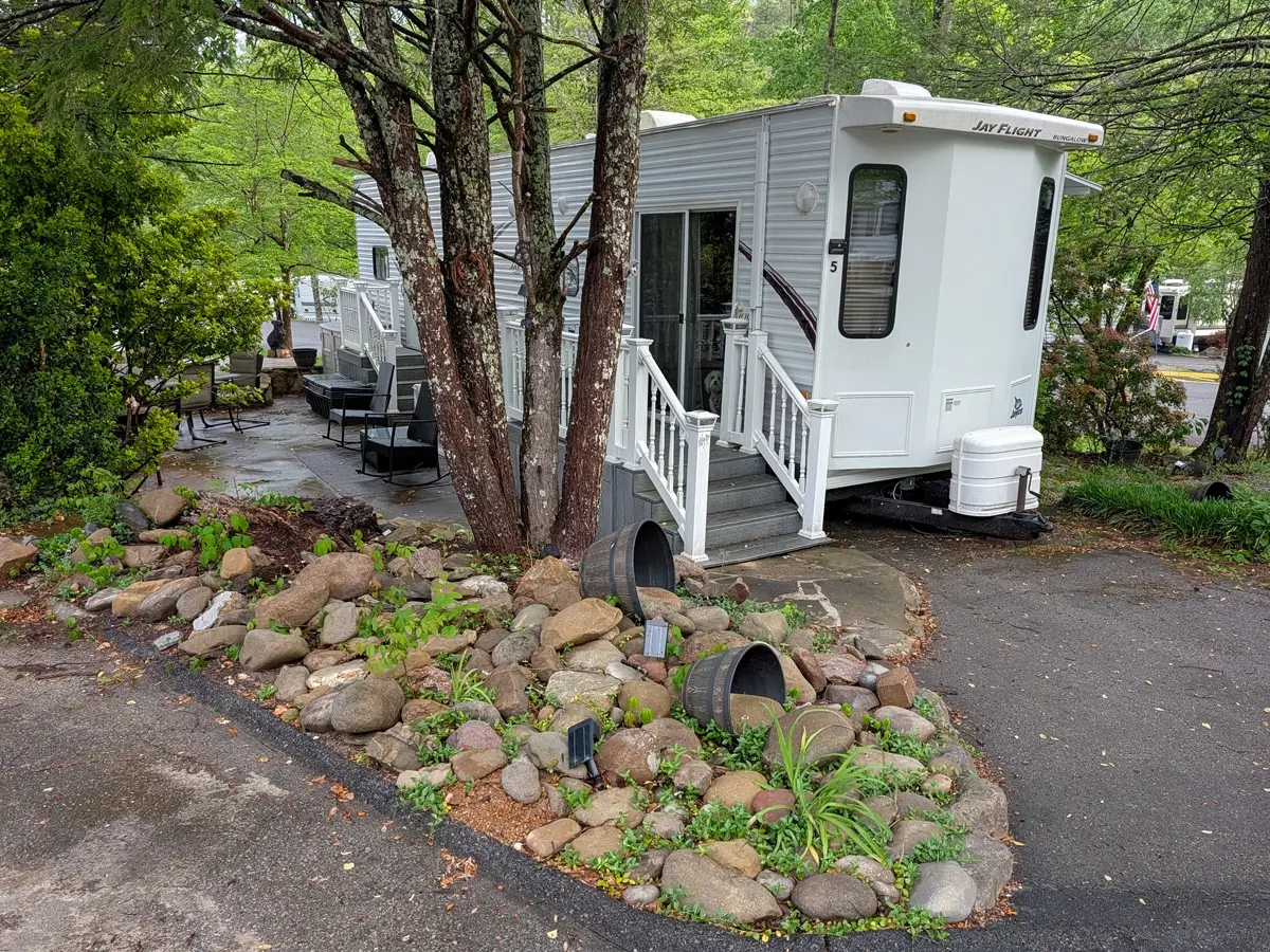 A white Jay Flight Bungalow travel trailer parked at a wooded campsite with a small stone-lined garden, used as a home base for a dog-friendly trip to the Smokies.