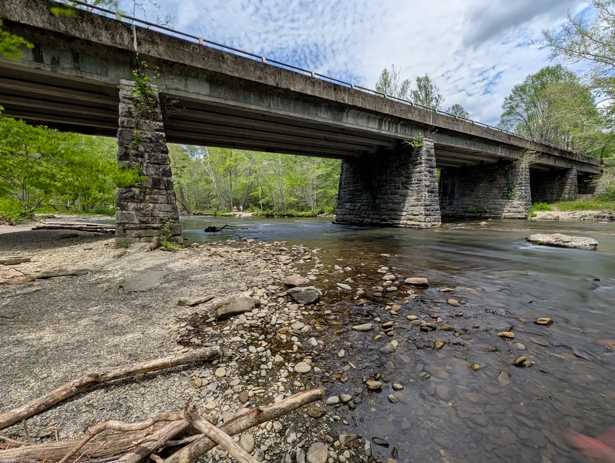 A large stone and concrete bridge spanning the Oconaluftee River, with a rocky shoreline in the foreground near the Great Smoky Mountains National Park visitor center.