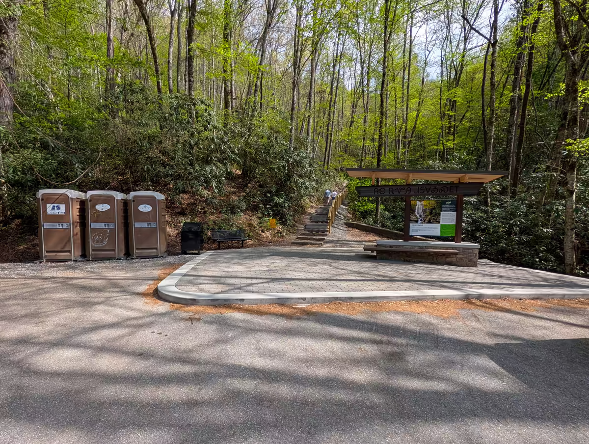 Mingo Falls Access Area The Mingo Falls NC trailhead area, featuring a paved parking spot, a row of portable toilets, and an information kiosk under a wooden pavilion, with a wooded path leading toward the falls.