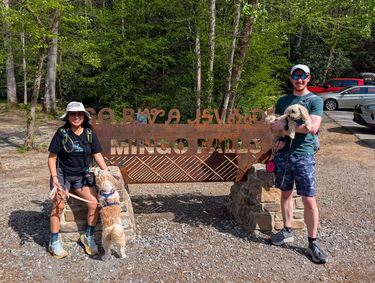 Mingo Falls Sign Sam and Minerva standing in front of a rustic, metal "Mingo Falls" sign at the trailhead, with the woman holding a small dog on a leash and the man holding another small dog in his arms.