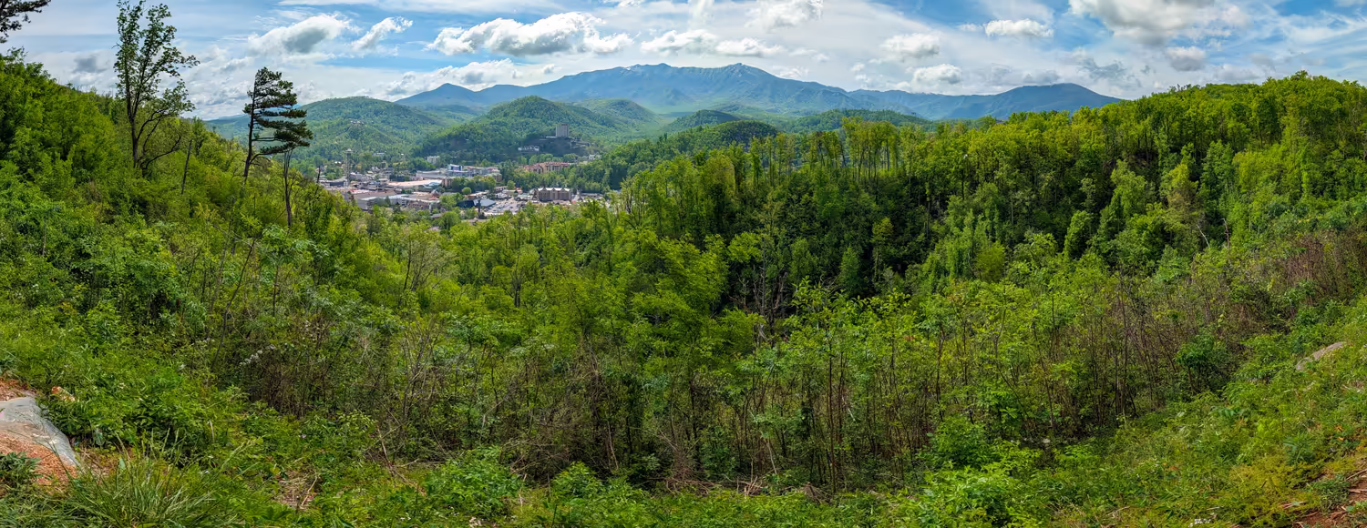 Panoramic overlook view of downtown Gatlinburg nestled in a lush green valley, surrounded by dense forest trees with the Great Smoky Mountains rising in the background under a cloudy sky.