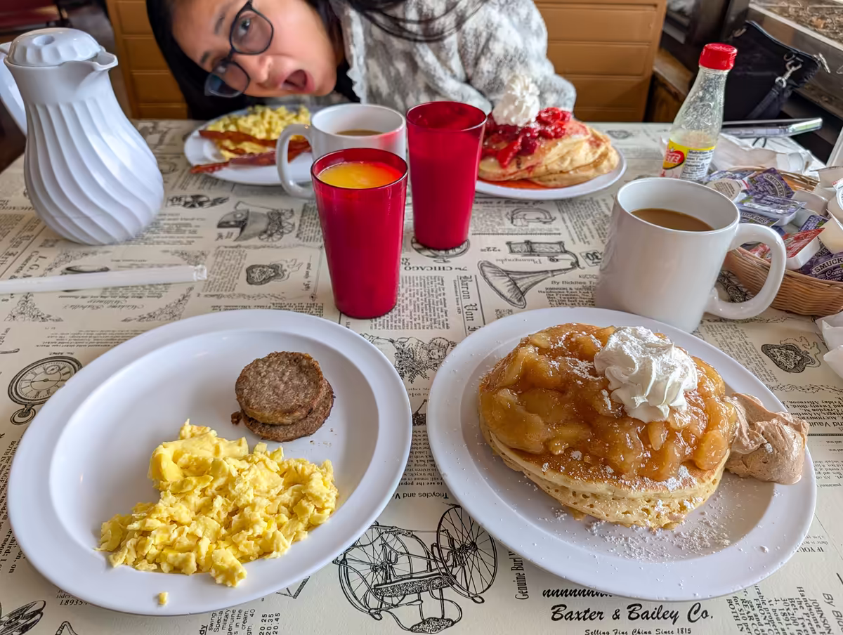 A hearty breakfast table spread at Little House of Pancakes in Gatlinburg, featuring stacks of pancakes topped with apples, strawberries, and whipped cream, alongside plates of scrambled eggs, sausage patties, and bacon.